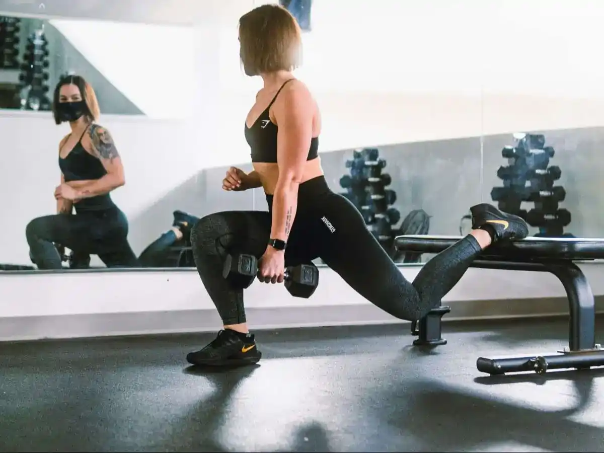 The image shows a woman performing a Bulgarian split squat in a gym setting. She is holding dumbbells in each hand while her left leg is positioned on a bench behind her and her right leg is bent at a 90-degree angle, with her foot firmly on the ground. The woman is wearing black workout leggings, a black sports bra, and black sneakers with a yellow Nike swoosh. A mirror on the wall reflects her image as well as part of the gym environment, including weight racks in the background. Another person, wearing a mask and dark workout clothes, is visible in the mirror, observing or possibly assisting her from a distance. The lighting creates a clear reflection of both individuals and highlights the workout space.