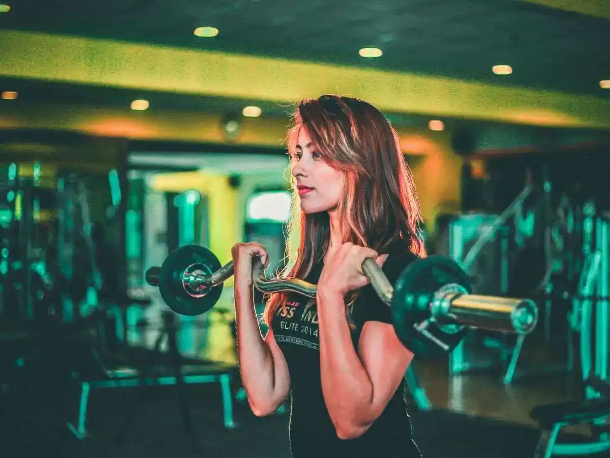 A woman is performing a barbell curl exercise inside a gym. She holds a barbell in front of her chest, with both hands gripping it firmly, lifting it toward her shoulders. Her long, reddish-brown hair is down, and she is wearing a black T-shirt. The gym environment is dimly lit, with overhead lights casting a warm glow. Behind her, various gym equipment, including benches and weight machines, can be seen in the background. The overall ambiance of the space is calm yet focused, enhancing the workout scene. The reflection of lights adds depth to the background.