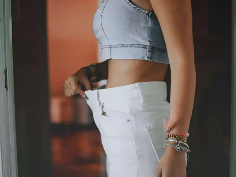 The image shows a woman standing sideways, wearing a light gray crop top and white pants that appear loose around her waist, suggesting weight loss or a change in size. She is holding the waistband of the pants with her left hand, pulling it away from her body to emphasize the extra space. Her arm, adorned with several bracelets in silver and gold tones, hangs naturally by her side. The background is softly blurred, with a warm, reddish-brown hue that contrasts with her outfit and brings focus to her torso and the loose pants. The lighting highlights her midriff and waistline, emphasizing the fit of the clothing. The scene gives an impression of a personal achievement in fitness or weight management.