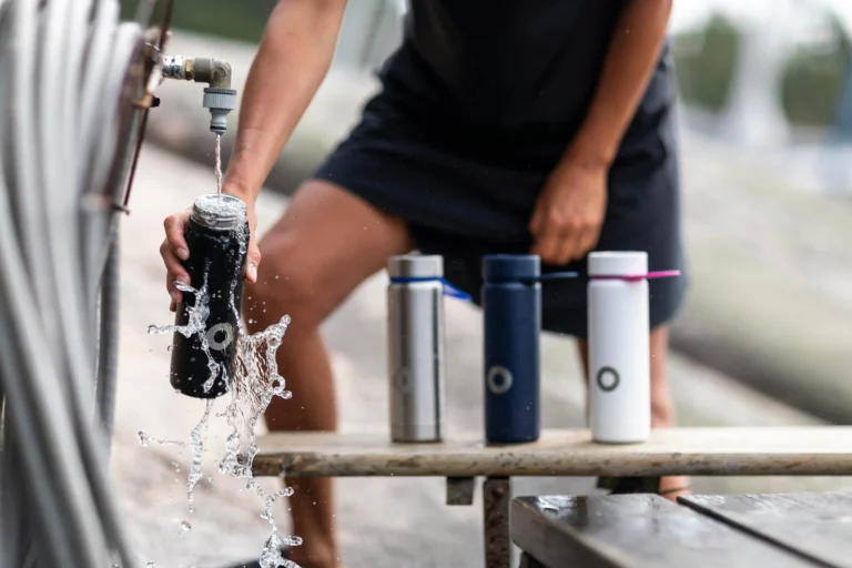 A person wearing a black outfit is filling a black water bottle from an outdoor faucet, with water splashing over the sides. The faucet is attached to a metal pipe with coiled hoses nearby. Three additional reusable bottles in silver, navy blue, and white are neatly lined up on a wooden bench beside the person. The white bottle has a pink straw attachment, while the others have a simple cap design. The background is blurred, showing an outdoor setting with natural light. The wooden bench appears weathered, supporting the bottles and adding a rustic touch. The overall scene emphasizes hydration, sustainability, and outdoor activity. 