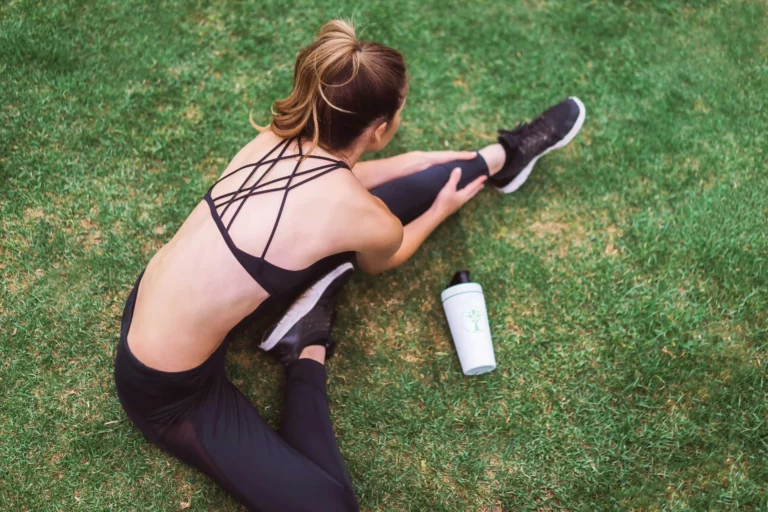 A woman with light skin and a toned physique is sitting on a grassy field, stretching her right leg while wearing a black sports bra with crisscross straps and black leggings. Her blonde hair is tied up in a high ponytail, and she is wearing black athletic shoes with white soles. Her right arm is extended forward, holding her right ankle, while her left hand supports her leg. A white and green shaker bottle with a tree logo is placed on the grass near her left side. The grass beneath her is lush and slightly uneven, with patches of lighter and darker green shades. The background is out of focus, emphasizing her stretching posture and the fitness-related theme. The natural lighting suggests an outdoor setting, possibly a park or an open field.